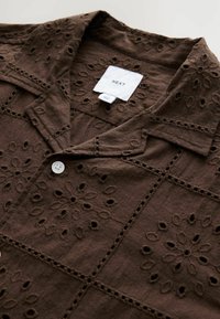 Chemise boutonnée en coton marron avec des découpes en broderie florale, un col et une étiquette portant la mention "NEXT" près de l'encolure.