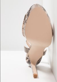 Chaussure à talon haut avec semelle beige et dessus imprimé serpent, positionnée semelle vers le haut contre une surface blanche à angles.