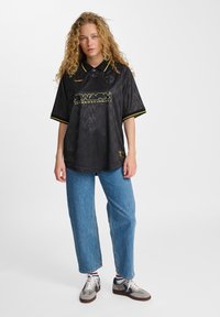 Μαύρο, κοντομάνικο πόλο μπλουζάκι με υφασμάτινη υφή. Διαθέτει κίτρινες λεπτομέρειες, λογότυπο και κλασικό γιακά. Συνδυάζεται με μπλε τζιν και αθλητικά παπούτσια.