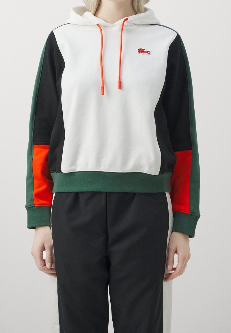 Personne portant un sweat à capuche en blocs de couleurs blanc, noir, vert et rouge avec des cordons orange, et un pantalon noir avec des bandes blanches sur les côtés.