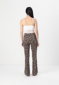 Bílý krajkový crop top s tenkými ramínky, doplněný o hnědé kalhoty s leopardím potiskem a rozšířenými nohavicemi, které ukazují hladkou strukturu a odvážný vzor.