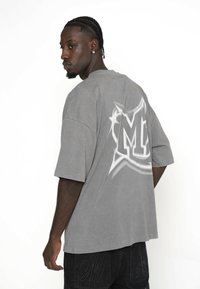 Szary oversize t-shirt z dużą białą grafiką na plecach. Krótkie rękawy, luźny krój i miękka faktura materiału. Zestawiony z ciemnoniebieskimi dżinsami.
