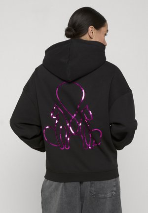 Person iført svart hettegenser med paljettpyntet rosa omriss av to katter som sitter rygg mot rygg, med halene sammenflettet til en hjerteform.