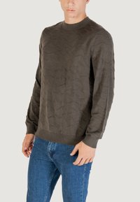 Dunkelgrüner strukturierter Pullover mit Wellenmuster, Rundhalsausschnitt und gerippten Bündchen. Kombiniert mit blauen Jeans.