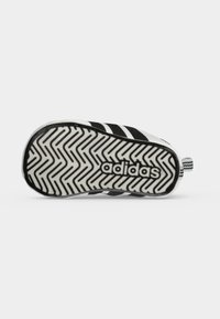 Suela de zapatilla Adidas en blanco y negro con patrón de espina de pescado y logo de Adidas en el centro sobre un fondo gris claro.