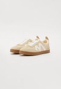 Sneakers atletiche beige con tomaia in rete, dettagli bianchi, colletto imbottito e suola in gomma. Presentano un'iconica logo a "M" sul lato.