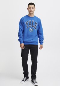 Felpa blu con la scritta "Yale" in grandi lettere bianche, accompagnata da un logo circolare e decorazioni a foglia di alloro. Abbinata a pantaloni neri e scarpe nere.