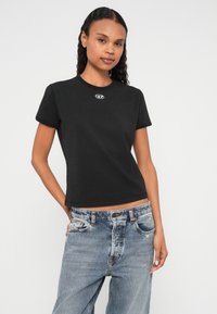 Melna kokvilnas T-krekls ar īsām piedurknēm un pieguļošu dizainu. Pie kakla neliels balts logotips. Kombinēts ar nodilušām zilām džinsa biksēm.