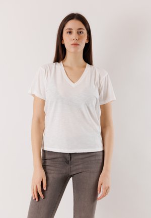 Frau mit langen braunen Haaren, die ein weißes V-Ausschnitt-T-Shirt und graue Jeans trägt, vor einem schlichten weißen Hintergrund stehend.