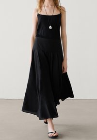 Sort maxi-nederdel med et tætsiddende, struktureret taljebånd og en flydende silhuet. Bæres med en sort tanktop og flade sorte sandaler.