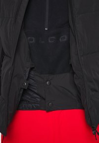 Svart vadderad jacka med dragkedja och knäppning, med ett mjukt inre fleece-lager. Inkluderar elastisk nederkant och en slät textur.