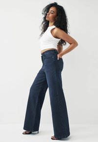 Højtaljede, brede jeans i mørkeblåt denim med en glat tekstur og minimal syning. Parret med en hvid ærmeløs top.