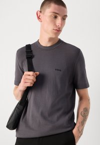 Szürke kötött, rövid ujjú póló texturált vállbetétekkel és diszkrét logóval. Fekete crossbody táskával párosítva.
