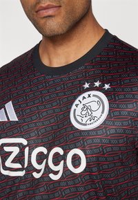 Černý fotbalový dres s grafickými červenými a šedými pruhy. Obsahuje znak Ajax, logo Ziggo a tři hvězdy nad emblémem. Kola s límcem.