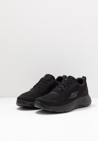 Schwarze Sportschuhe mit einem atmungsaktiven Mesh-Obermaterial, runder Zehenpartie, flexibler Sohle und strukturierten Akzenten sowie einem Logo an der Seite.