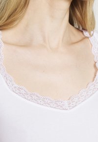 Bílý top s jemným růžovým krajkovým výstřihem. Krajka má zvlněný okraj s květinovými vzory, které doplňují hladkou texturu látky.