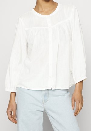 Persona con blusa blanca abotonada de corte amplio y mangas largas, y vaqueros azul claro, de pie contra un fondo liso.