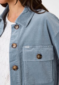 Nahaufnahme einer Person, die eine hellblaue Cordjacke mit großen braunen Knöpfen und einer Vorderasche trägt, die mit "Garcia" beschriftet ist.