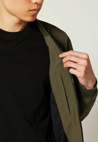 Olivgrüne Bomberjacke mit einem Reißverschluss vorne, gerippten Bündchen und leichtem Material. Darunter befindet sich ein schwarzes T-Shirt.