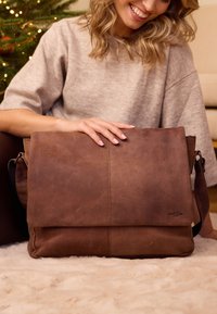 Bolsa de mensajero de cuero marrón con una solapa plegada, que presenta una textura suave, costuras visibles y una correa ajustable.