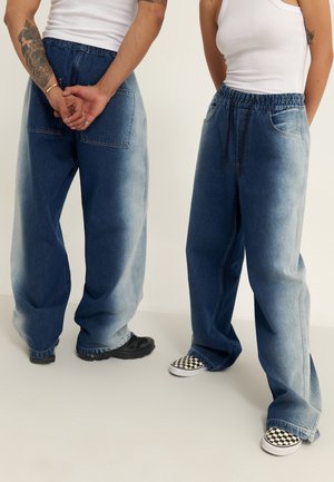 Dos personas con camisetas sin mangas blancas y pantalones vaqueros holgados de dos tonos azules, una con brazos tatuados y zapatos slip-on de cuadros.
