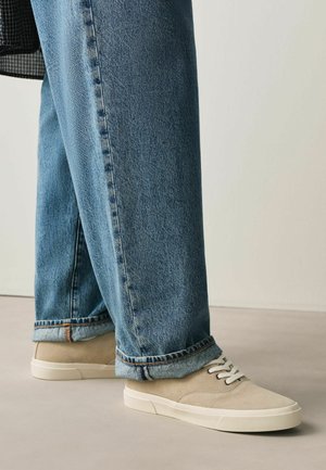 Bēšas zamšādas sporta kurpes ar baltām zolēm un šņorēm, valkātas kopā ar gurnu salocītām gaiši zilām taisnām džinsa biksēm uz neitrālas grīdas.