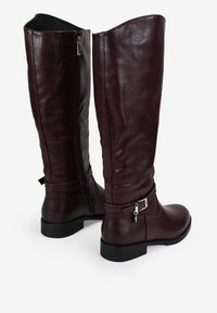 Botas de cuero burdeos hasta la rodilla con una textura suave, cremallera lateral y detalle de tira en el tobillo con un hebilla metálica. Suelas planas.