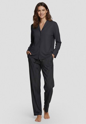 Woman standing barefoot wearing a black long-sleeve buttoned shirt and black pinstriped pants with hands in pockets.