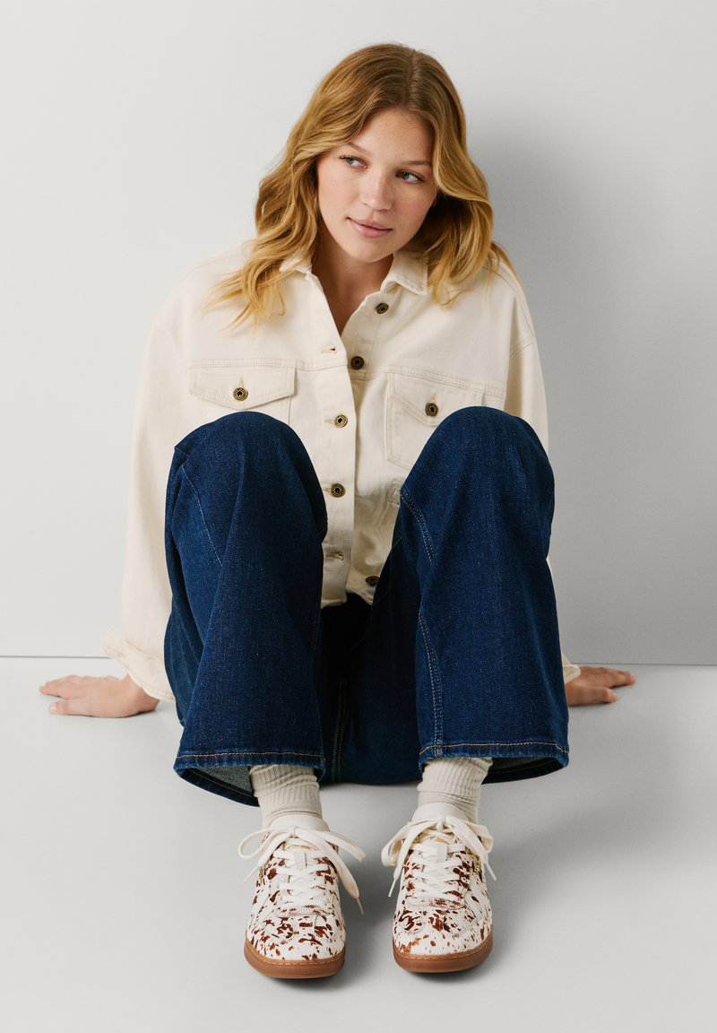 Νεαρή γυναίκα με ξανθά μαλλιά κάθεται δίπλα σε τοίχο, φοράει κρεμ σακάκι, μπλε τζιν, κρεμ κάλτσες και λευκά αθλητικά παπούτσια με καφέ σημεία.