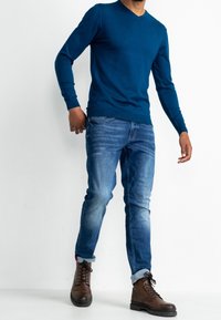 Blauer Pullover mit V-Ausschnitt, eng anliegende hellblaue Jeans und braune Schnürstiefel mit strukturierter Oberfläche. Jeans sind an den Bündchen leicht umgeschlagen.