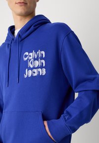 Blauwe katoenen hoodie met een voorste kangoeroe zak, een capuchon met koord en een geborduurd "Calvin Klein Jeans" logo in het wit.