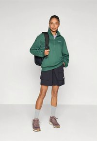 Donna che indossa un felpa a zip verde, pantaloni cargo neri, calzini grigi, scarpe da trekking color malva, e porta uno zaino nero su una spalla.