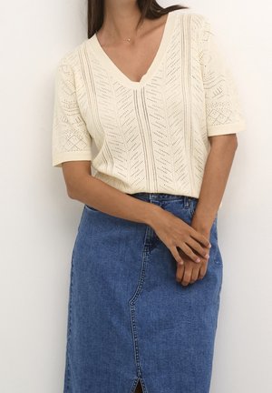 Kobieta w kremowym, krótkim swetrze z dekoltem w serek i niebieskiej dżinsowej spódnicy z przednim rozcięciem, stoi ze złożonymi rękami.