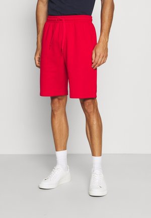 Male wearing bright red drawstring shorts, white socks, and white sneakers, standing against a plain light-colored background.