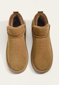 Para brązowych zamszowych kapci z miękkim futrem wewnątrz i gumową podeszwą, widziana z góry na jasnym tle.