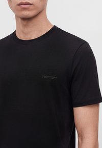 Άνδρας που φοράει απλό μαύρο t-shirt Armani Exchange με μικρό λογότυπο στο αριστερό στήθος, κομμένο κάτω από τη μύτη και πάνω από τον αγκώνα.