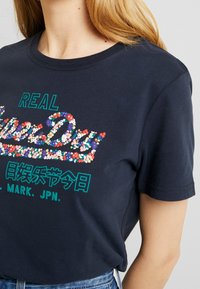 Marinblå bomulls t-shirt med färgglada blomster-sekvensgrafiker med "REAL SuperDry" och japansk text. Kort ärm och design med rund hals.