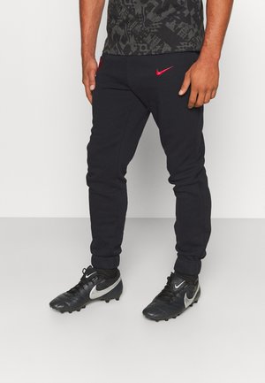 Homme portant un pantalon de survêtement Nike noir avec un logo rouge et des crampons Nike noirs avec le swoosh blanc, debout devant un fond clair uni.