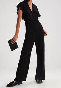 Mono negro con mangas cortas con volantes, diseño de cuello en V y corte de pata ancha, combinado con un bolso de mano negro y zapatos planos. Textura suave.