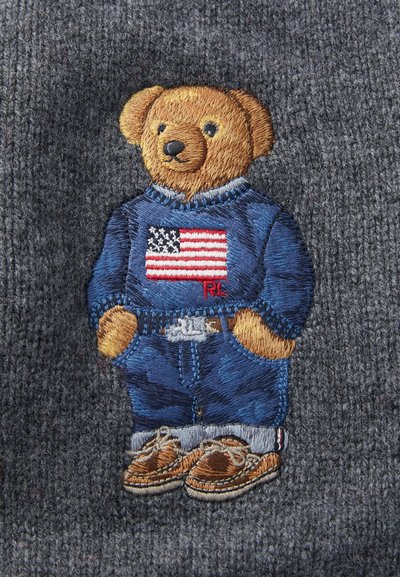 Izšūts lācis zilos džemperī ar Amerikas karogu, džinsu kopēšanās un brūnās kurpes, uz teksturēta pelēka fona.