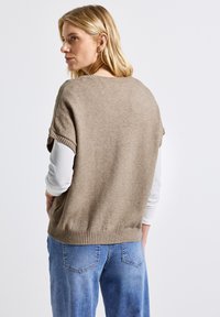 Khaki gebreide trui met korte mouwen, geribde zoom, gedragen over een witte longsleeve. Heeft een relaxed fit en een zachte textuur.