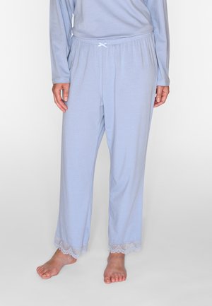 Persona che indossa pantaloni del pigiama azzurri chiaro con bordatura in pizzo alle caviglie e una maglia a maniche lunghe abbinata, in piedi scalza su un pavimento bianco.