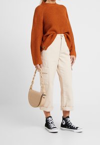 Person som bär en roströd stickad tröja, uppvikta ljusbeige cargobyxor, svarta höga sneakers och håller en beige handväska med flätat handtag.