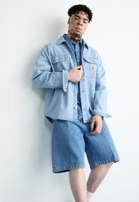Lyseblå denimjakke med brystlommer, parret med en matchende denimskjorte og shorts. Har en afslappet pasform og diskrete syninger.