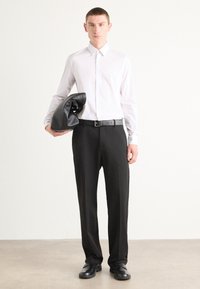 Weißes Hemd mit Knöpfen, schwarze Hose, schwarzer Gürtel. Hält eine zerknitterte schwarze Ledertasche. Glatter, taillierter Stoff, minimalistisches Design.