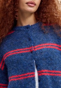 Vrouw met krullend rood haar, gekleed in een blauwe gebreide cardigan met horizontale rode strepen en een witte top eronder, weergegeven van nek tot taille.