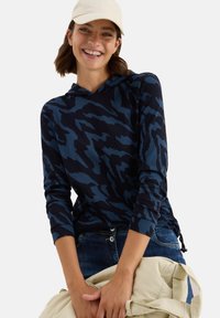 Glunderende vrouw in een blauw-zwarte, met een patroon bedrukte hoodie en een beige pet, met een beige tas in haar hand, gekleed in blauwe jeans voor een witte achtergrond.