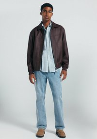 Braune Lederjacke mit einem Kragen, getragen über einem hellblauen karierten Hemd, kombiniert mit hellblauen Jeans und braunen Slipschuhen.