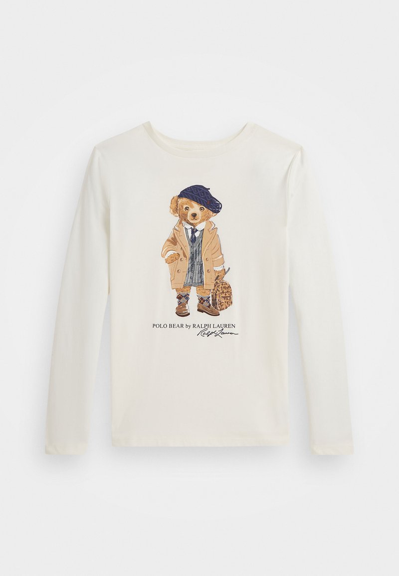 Κρέμα μπλούζα μακρύ μανίκι με απεικόνιση ενός αρκούδου σε καφέ παλτό, μπλε μπερέ και ανοιγμένο γιλέκο, κρατώντας μια τσάντα με λεοπάρ σχεδιασμό.