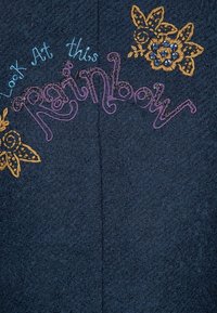 Un tessuto blu scuro presenta il testo ricamato "Guarda questo Arcobaleno" in rosa, con design floreali in giallo e abbellimenti blu.